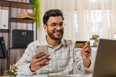 Mutlu Hintli işadamı banka kartı ve akıllı telefonu ev ofisteki online alışveriş ödemeleri için kullanıyor. İş anlaşması. Gülümseyen adam cep telefonu E-bankacılık uygulama servisiyle serbest alım yapıyor..