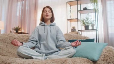 Sakin ol, rahatla, iç denge. Genç kadın derin nefes alıyor Mudra jesti ile, gözleri kapalı meditasyon yapıyor konsantre düşüncelerle, huzurlu zihinle. Kanepedeki kız lotus pozisyonunda sakin görünüyor.
