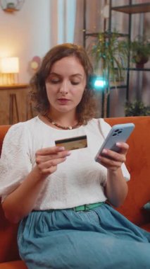 Para transfer ederken kredi kartı akıllı telefonu kullanan gülümseyen genç bir kadın, kapalı bir evde çevrimiçi alışveriş nakitsiz yemek siparişi veriyor. Kanepede oturan mutlu kız. Dikey