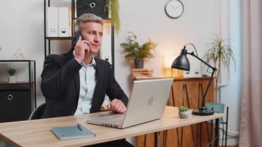 Genç işadamı bilgisayarını kullanıyor ve cep telefonuyla müşteriye cevap veriyor. Resmi takım elbiseli beyaz adam uzaktan kumanda işinde çalışıyor, ofis masasında konuşuyor..