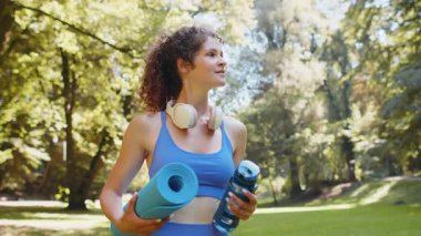 Elinde hasırla genç ve mutlu sporcu kadın parktaki açık hava eğitimine gidiyor. Aktif giyimli, kulaklık takan ve su şişesi kullanan iyimser gülümseyen kız jimnastik eğitimine gidiyor..