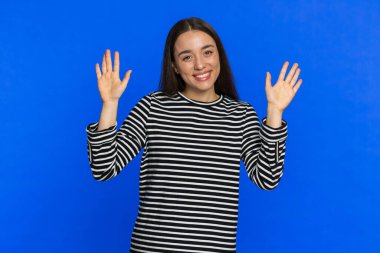 Kameraya dostça gülümseyen mutlu beyaz kadın, iki elini sallayarak selam veriyor ya da davetkar bir yüz ifadesiyle veda ediyor. Güzel kız mavi stüdyo arka planında izole edilmiş.
