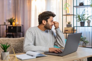 Genç Hintli adam dizüstü bilgisayarda çalışıyor, yorgun görünüyor, evdeki oturma odasındaki kanepede otururken gözlerini ovuşturuyor. İspanyol bir işkolik, görevine devam ediyor, bitkin görünüyor..