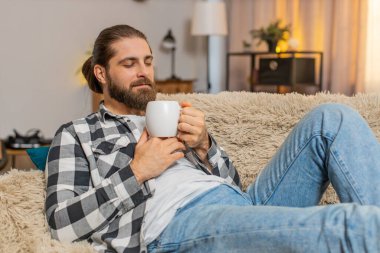 Genç adam evde dinleniyor, elinde sıcak bir fincan kahve ya da çay tutarak nazikçe sırıtıyor ve omuzlarını gevşetiyor. Kanepedeki beyaz adam dinlendirici bir sessizlik hissediyor, yoğun görevlerden ve gürültüden kısa bir ara veriyor..