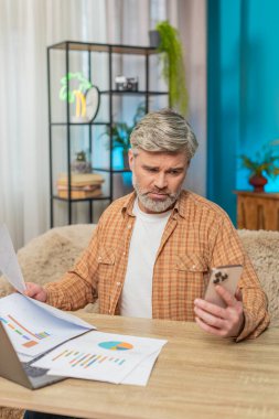 Beyaz, olgun bir adam evde, dizüstü bilgisayar ve akıllı telefon kullanıyor, mali raporları inceliyor. Orta yaşlı bir adam yorgun görünüyor, gözlerini ovuşturuyor ve finansal sorunlara özenle çözüm bulmaya çalışıyor.
