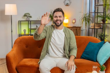 Kameraya gülümseyen, el sallayan, el sallayan, evdeki oturma odasındaki kanepede otururken selamlaşan mutlu beyaz adam. Mutlu sakallı adam evde video kaydediyor..