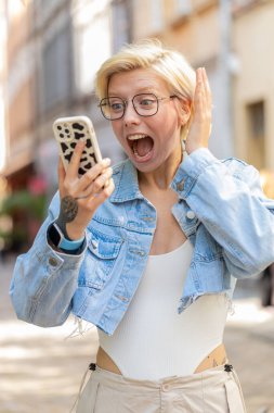 Kafkasyalı genç bir kadın cep telefonuyla kutlama yapıyor iyi haber kazanıyor, piyango büyük ikramiyesi kazanıyor, açık havada dağıtıyor. Şehir caddesinde zıplayan mutlu sarışın bayan turist. Kasaba yaşam tarzları.