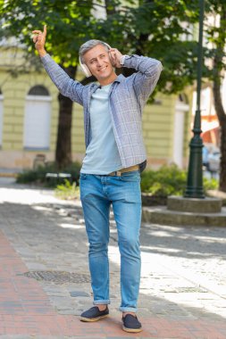 Neşeli, aşırı mutlu, kır saçlı, kablosuz kulaklıklı adam disko müziği dinliyor. Şehir parkında günlük giysiler içinde yürüyen neşeli bir adam. Kasaba yaşam tarzları. Dikey.