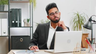 Modern ofis çalışma masasındaki mutlu, sakallı Hintli işadamının portresi başını kameraya çevir. Resmi takım elbiseli bir erkek Hindu. Yakışıklı Uzaktan kumandalı adam dizüstü bilgisayarla çalışıyor