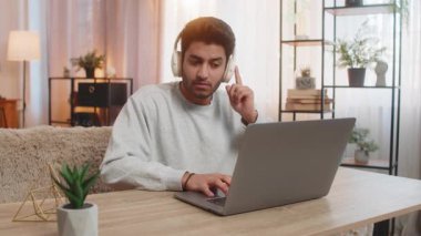 Kablosuz kulaklık takan genç Hintli adam dizüstü bilgisayarda çalışıyor, sesli kitap dinlerken odaklanmış görünüyor. İspanyol adam evdeki oturma odasındaki koltukta oturuyor, göreviyle çok meşgul..