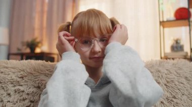 Genç bir kız evde düşünceli ve odaklanmış bir ifadeyle gözlük takıyor. Kafkasyalı çocuk kanepede oturuyor ve rahat oturma odasında günbatımında sakin ve pratik bir tavırla göz giysisini ayarlıyor.