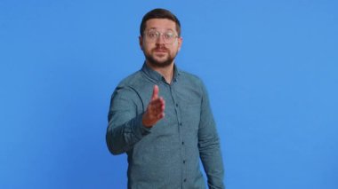 Dost canlısı, orta yaşlı, gözlüklü, kameraya uzanan, el sıkışan, nazik bir gülümsemeyle selamlayan, davetli, dayanışma gösteren, mavi arka planda güven konsepti olan orta yaşlı bir adam.