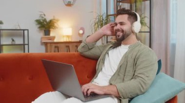 Happy, kablosuz kulaklıklı, rahat, Kafkasyalı genç adam kanepede oturup dizüstü bilgisayar ağı üzerinde çalışırken disko müziği dinlemeyi seçiyor. Hafta sonu evde eğlenen yakışıklı bir adam.