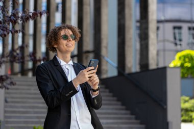 Kafkasyalı mutlu iş adamı akıllı telefon daktilosu kullanıyor e-posta mesajlarına cevap veriyor açık havada internetteki mobil uygulamalarla sohbet ediyor. Şehir merkezinde takım elbiseli kıvırcık saçlı yakışıklı bir adam. İş adamları