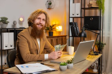 Beyaz saçlı mutlu iş adamı nakit sayıyor ve ofis bilgisayarını kullanıyor. Başarılı bir serbest çalışan ofise bir tomar para getiriyor, temettüleri hesaplıyor nakit para kazancını hesaplıyor.