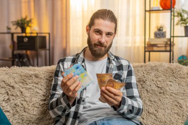 Genç adam evde taze para nakit banknotlarını teftiş ediyor, her avroyu belli bir odak noktasıyla dikkatlice ayırıyor. Kanepede oturan beyaz adam memnun görünüyor. Bütçe ilerlemesi ve ihtiyaçları için düzenli bir gelir sağlıyor.