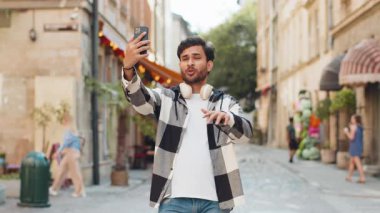 Neşeli Hintli blog yazarı akıllı telefondan selfie çekiyor, aile dostlarıyla internet üzerinden video görüşmesi yapıyor sosyal medya vlog 'u için hikayeler kaydediyor, el sallıyor. Şehir sokağındaki İspanyol adam..