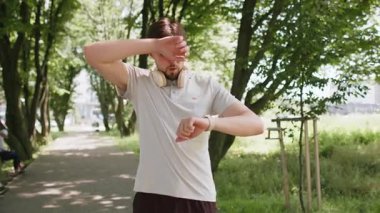 Yoğun kardiyo eğitimi. Genç, aktif bir erkek koşucu parkta koşuyor ve akıllı adımlara bakıyor, yoruluyor ve yavaşlıyor. Sporcu, dışarıda ağaçlarla kalp atışlarını kontrol ediyor..
