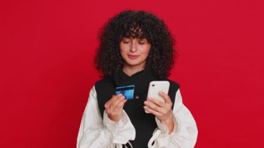 Kredi kartı ve akıllı telefon kullanarak internet üzerinden alışveriş yapan, yemek siparişi veren kıvırcık saçlı mutlu beyaz kadın. Kırmızı arkaplanda izole edilmiş kız