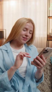 Genç bir kadın favori restoran yemeği ve içeceklerini seçmek için telefon uygulamasını kullanarak evde oturuyor ve hızlı ödemeyi sonuçlandırıyor. Kanepede oturan beyaz kız gülümsüyor, zahmetsiz yemek dağıtım sürecinden tatmin oluyor.