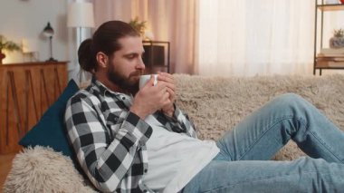 Genç adam evde dinleniyor, elinde sıcak bir fincan kahve ya da çay tutarak nazikçe sırıtıyor ve omuzlarını gevşetiyor. Kanepedeki beyaz adam dinlendirici bir sessizlik hissediyor, yoğun görevlerden ve gürültüden kısa bir ara veriyor..