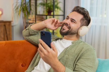 Kablosuz kulaklıklı, akıllı telefonlu mutlu genç adam evinde kanepeye uzanmış, en sevilen enerjik disko dans müziğini dinliyor. Hafta sonu eğlence aktiviteleri.