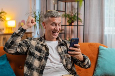 Heyecanlı genç adam elinde akıllı telefon tutarak online bahis oyununun kazanmasıyla hayrete düşen iyi haberleri okuyor. Kanepede oturan mutlu erkek. Evdeki zaferden çok memnun..