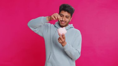 Kumbarayı elinde tutan mutlu Hintli genç adam bozuk para bırakarak para tasarrufu yapıyor, gelecekteki harcamaları ve finansal güvenliği planlıyor. Gülümseyen Arap adam birikimlerine yatırım yapıyor, pembe arka planda izole edilmiş.