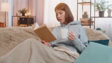 Rahat koltukta bir fincan sıcak çikolata, kakao ya da çay içerken ilginç bir kitap okuyarak rahatlayan genç kadın edebiyatın tadını çıkarıyor. Evdeki oturma odasında sıcak bir battaniyeye sarılı mutlu kız..