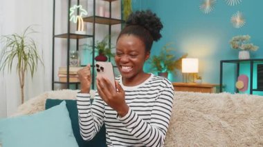 Afro-Amerikalı kadın akıllı telefondan iyi haberleri kutluyor. Evde kanepede oturmak, neşeli bir ifade, mutlu duygular, mesaj almak ya da piyangoyu kazanmak, kapalı mekan atmosferini meşgul etmek, heyecanlı ruh hali.
