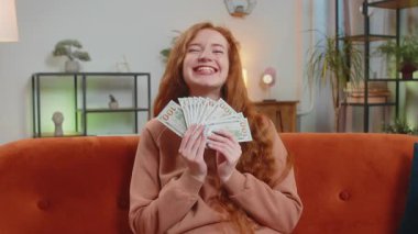 Başarılı, genç, beyaz, beyaz bir kadın elinde bir dolarlık banknot tutuyor ve evdeki kanepede oturuyor. Mutlu yetişkin kız çevrimiçi piyango oyunu kazanıyor tatil planlama bütçe hesaplama konsepti.