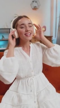 Kanepede oturmuş, kablosuz kulaklıkla müzik dinleyen mutlu genç bir kadın. Beyaz elbiseli neşeli yetişkin kız evdeki oturma odasında müzik eşliğinde dans ediyor. Dans müziğinin tadını çıkarıyorum. Dikey