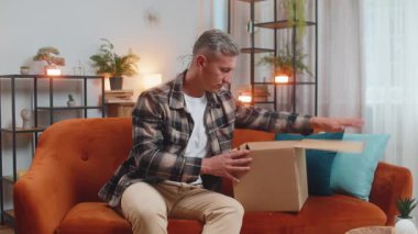 Evdeki kanepede oturan mutlu genç adam teslimat paketini açıyor. Gülümseyen memnun erkek alışverişci çevrimiçi dükkan müşterisi karton kutu açıyor hızlı posta yoluyla satın alma hediyesi alıyor.