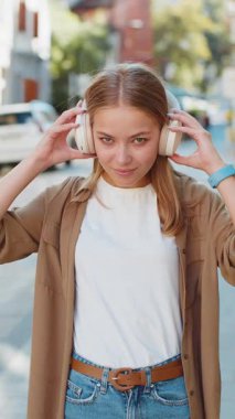 Müzik dinleyen mutlu beyaz kadın portresi. Kulaklıklarını çıkarıyor, kameraya bakıyor, rahatlıyor ve dışarıda iyi haberler veriyor. Şehir parkındaki bayan kız. Dikey