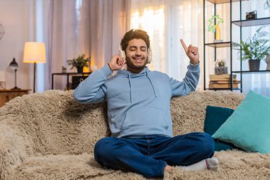 Kanepede oturan Hintli genç adam kulaklıkla müzik dinliyor ritimle dans ediyor rahatlıyor ve gevşiyor. İspanyol adam enerjiyle hareket etmeyi ve favori parçalarda neşe bulmayı seviyor..