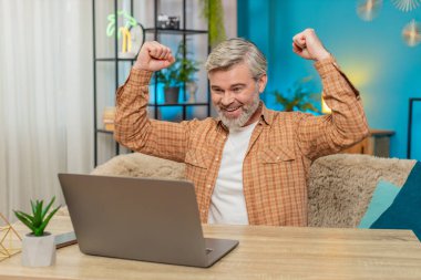 Laptop kullanan mutlu beyaz adam önemli bir e-posta mesajına ulaşıyor. İyi haber. Tatmin olmuş orta yaşlı bir adam evinde oturmuş sevinçle bağırıyor, oyunu kazandıktan sonra ellerini kaldırıyor.