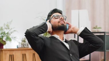 Mutlu gülümseyen Hintli işadamı kulaklıkla müzik dinliyor dinleniyor ofis yerinde çalıştıktan sonra sandalyeye yaslanıyor. Hindu serbest çalışan resmi takım elbiseli erkek ellerini başının arkasına koy.