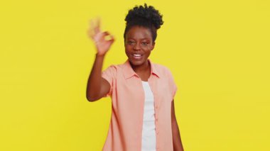 Afro-Amerikalı bir kadın tamam jest yapar, enerjiyi hisseder, olumlu geri bildirim yapar, onaylar, zaferi kutlar. Sarı arka plandaki siyah kız kendine güveni yayıyor, pozitif tavır sergiliyor