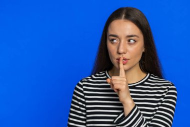 Sessiz olun lütfen. Beyaz kadın işaret parmağını dudaklarına bastırıyor. Sessiz jest işareti yapıyor. Sırları söyleme, dedikodu yapma, mahremiyetten bahsetme. Mavi arka planda izole edilmiş bir kız. Kopyalama alanı