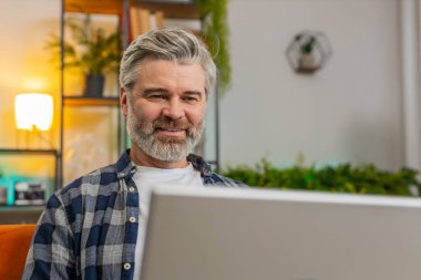 Gülümseyen sakallı, olgun bir adam dizüstü bilgisayarda yazı yazıyor. İnternette geziniyor. Kanepede tek başına oturan uygulamaları kullanıyor. Orta yaşlı, yaşlı, beyaz bir adam bilgisayarda çalışıyor. Oturma odasında ekrana bakıyor.