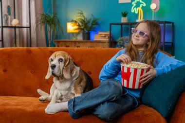Üç boyutlu gözlüklü, patlamış mısır yiyen ve ilginç TV dizileri, spor oyunları, filmler, çevrimiçi film içerikleri izleyen heyecanlı genç kız. Çocuk, evdeki oturma odasında kanepede av köpeğinin yanında oturuyor..