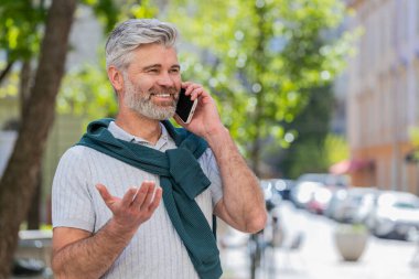 Sakallı, olgun bir turist akıllı telefonla konuşuyor. Şehir caddesinde beklenmedik bir şekilde iyi haber dedikodusu yapıyor. Şehir yaşam tarzları açık havada.