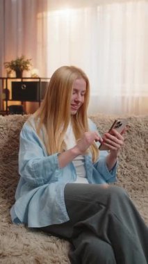 Genç kadın evde rahatlıyor, akıllı telefona bakıyor ve olumlu gelişmeleri büyük bir gülümsemeyle kutluyor. Kanepedeki beyaz kız heyecanla sıçrıyor, sıcak akşam güneşi altında muzaffer bir jest sergiliyor..