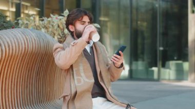 Mutlu Hintli genç adam sabah kahvesinin tadını çıkarıyor ve akıllı telefon kullanarak dışarı mesaj gönderiyor. Dinleniyorum, ara veriyorum. Şehir merkezindeki bankta oturan adam kahve içiyor..