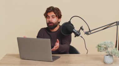 Mutlu blogcu adam podcast yapıyor, atölye veriyor, webinar yapıyor. Genç Hintli erkek mikrofon ve dizüstü bilgisayarda konuşuyor, video görüşmesi yapıyor. Bej arka planda izole edilmiş yakışıklı Hindu adam..