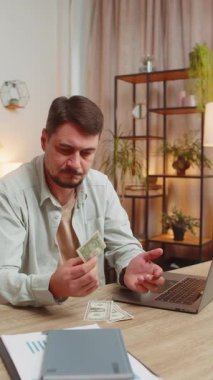 Kafkasyalı serbest çalışan bir adam dolar sayıyor. Yetersiz miktarda para. Finansal kriz. İflas. Yoksulluk ve yoksulluk. Ev ofis masasında dizüstü bilgisayarlı üzgün bir işadamı oturuyor. Dikey.