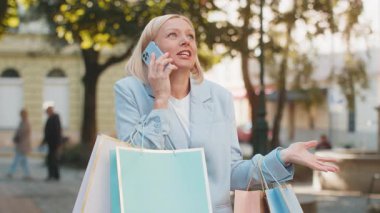 Mutlu beyaz iş kadını portresi gülümseyen şehir caddesinde elinde paketlerle durup telefonda konuşurken arkadaşına indirimlerden, alışveriş konseptinden bahsediyor..