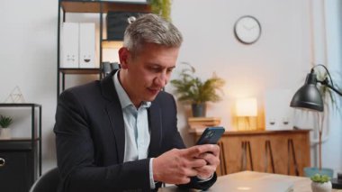 Beyaz adamın başarısını kutlarken dahili ofisteki çalışma masasında otururken akıllı telefondan harika haberler okuyor. Gülümseyen işadamı güzel bir tebliğden memnun, evet işareti gösteriyor..
