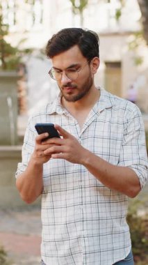 Gülümseyen Hintli genç adam akıllı telefon kullanıyor kısa mesajlar yazıyor internette geziniyor, işi bitiriyor, dışarıdaki kameralara bakıyor. Dışarıdaki şehir sokağında duran İspanyol turist. Kasaba yaşam tarzı