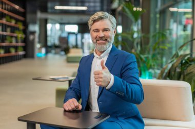 Gibi. Takım elbiseli mutlu, orta yaşlı bir işadamı kameraya bakıp olumlu bir işaret veriyor. Modern ofis lobisinde oturmuş olumlu bir izlenim veriyor. İş insanları.